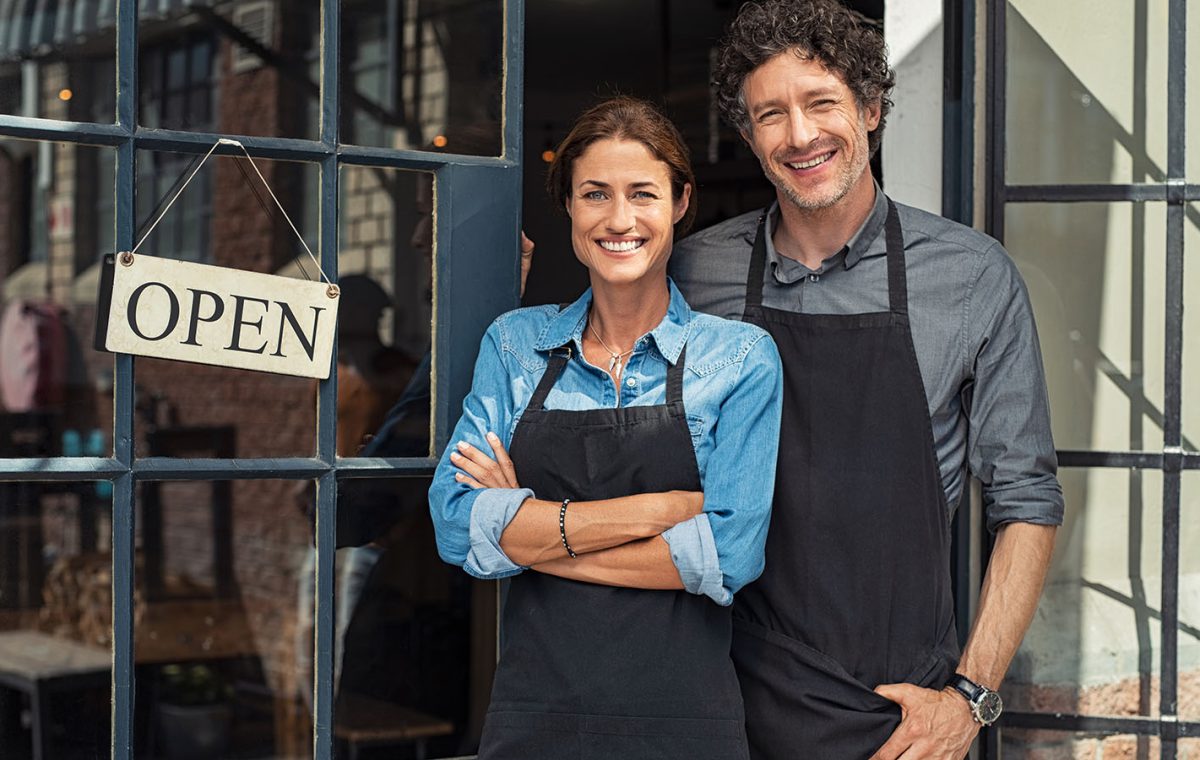 Two cheerful small business owners smiling and looking at camera while standing at entrance door. Happy mature man and mid woman at entrance of newly opened restaurant with open sign board. Smiling couple welcoming customers to small business shop.
